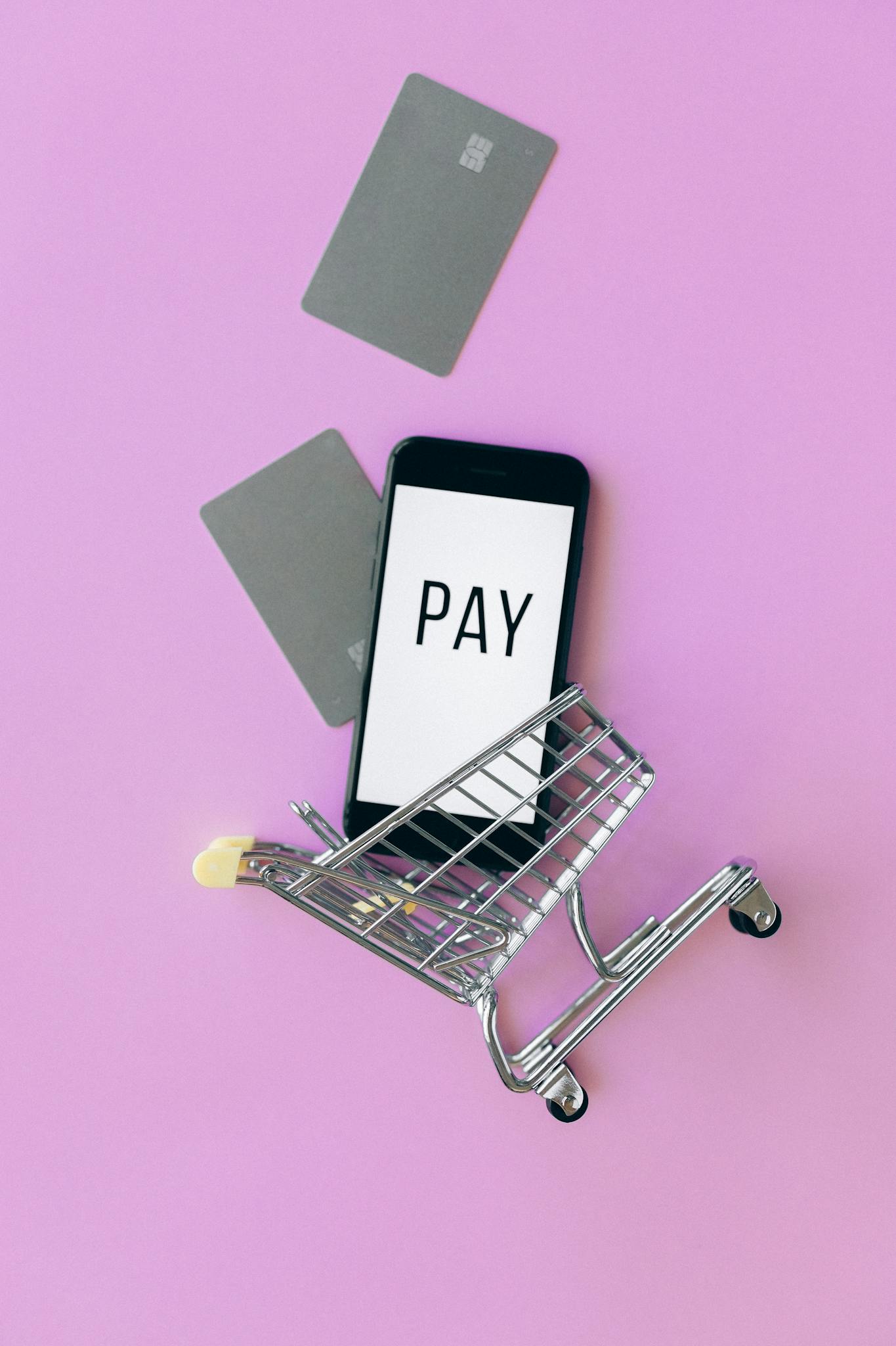 Top view of a smartphone in a shopping cart with credit cards on a purple background.